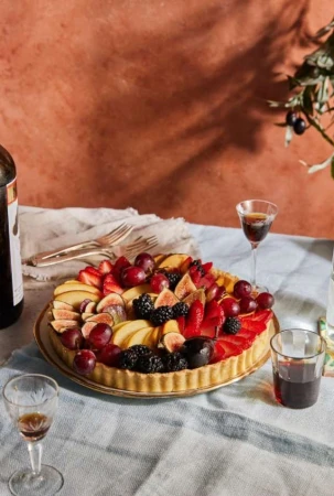 Fruit tart topped with figs, strawberries, and grapes on a table with wine glasses and a textured wall background
