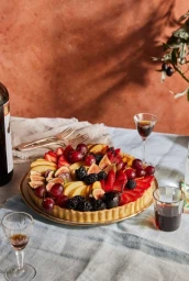Fruit tart topped with figs, strawberries, and grapes on a table with wine glasses and a textured wall background