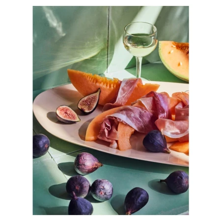 Proscuitto and melon slices arranged on a platter with fresh figs and a glass of white wine on a green tiled surface