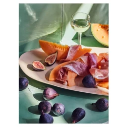 Proscuitto and melon slices arranged on a platter with fresh figs and a glass of white wine on a green tiled surface
