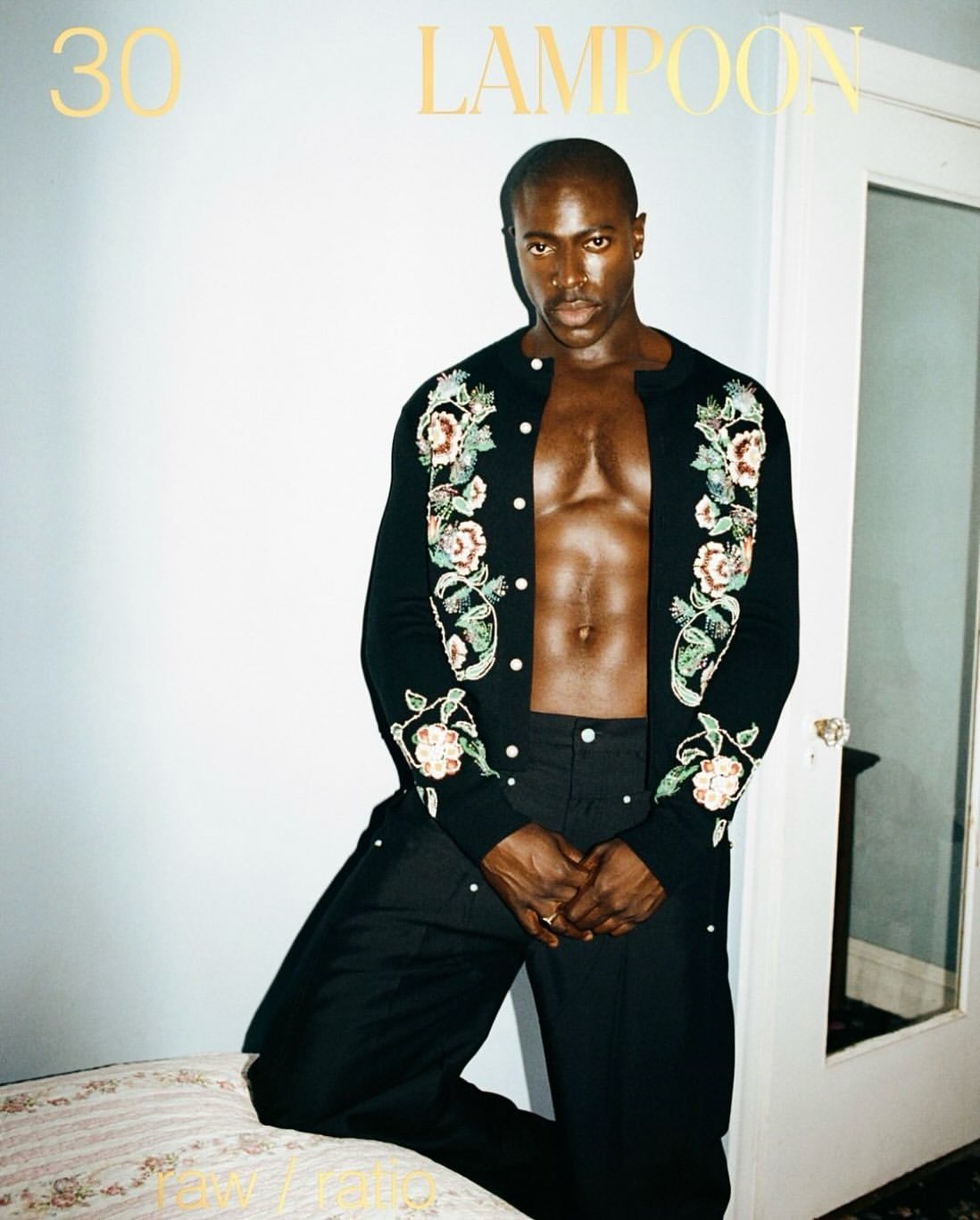 An African-American man poses indoors wearing an open black floral cardigan and black pants, against a white wall with a door.