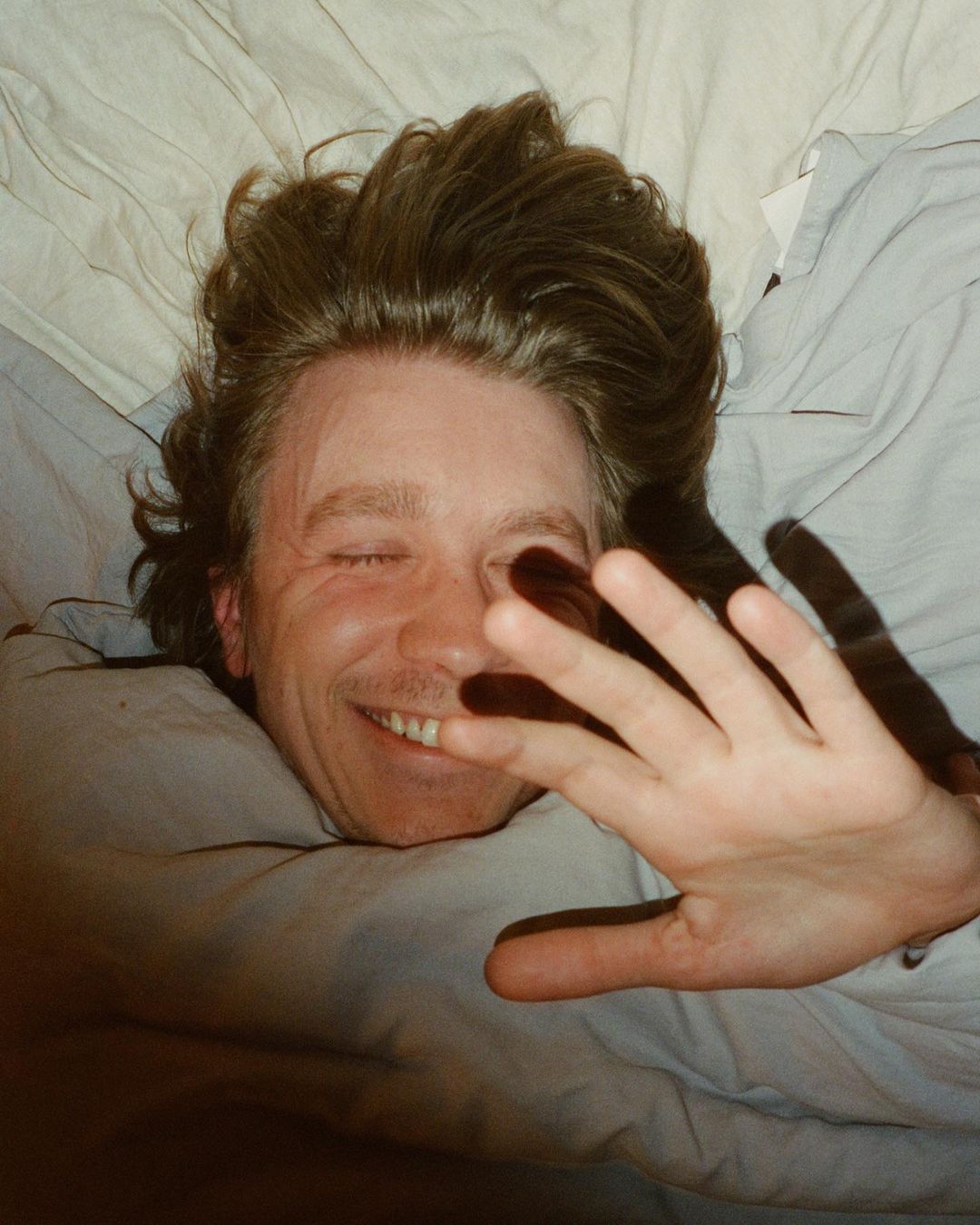 A man lies in a bed with white sheets and pillows, smiling with his eyes closed, and his hand partially obscuring his face.