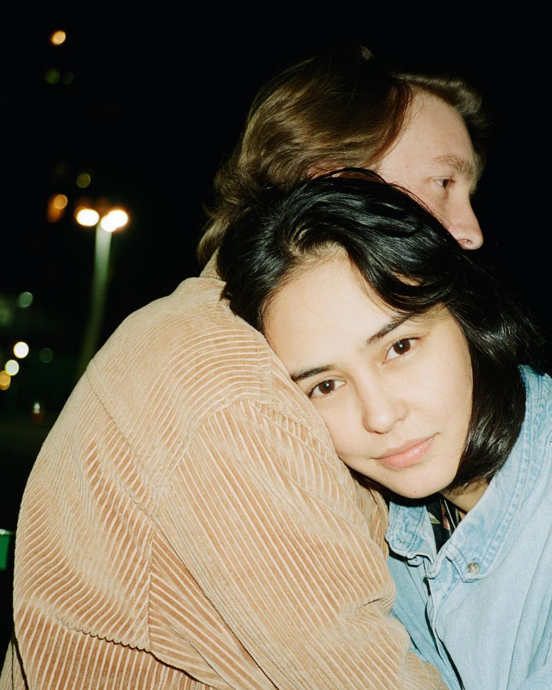 Portrait of a hugging couple outdoors at night. The woman faces the camera, leaning on the man's shoulder in the darkness with street lights.