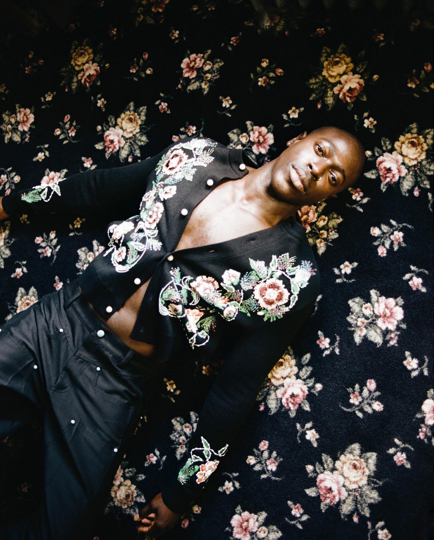 An adult male wearing a black floral-embroidered cardigan and pants is lying on a floral patterned surface in an indoor environment.