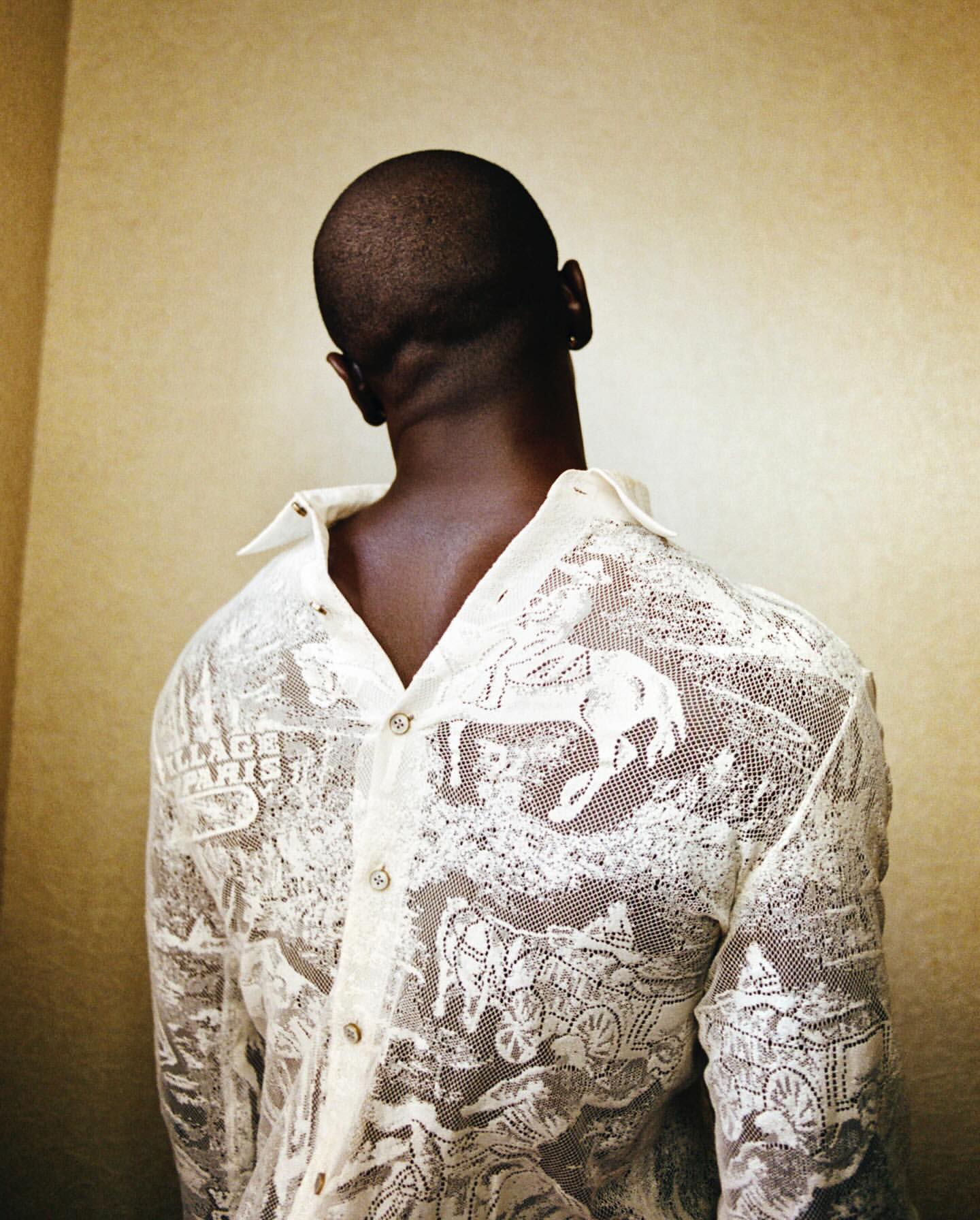 Back view of an adult Black man wearing a sheer white button-down shirt with vintage horse and buggy design against a neutral wall.