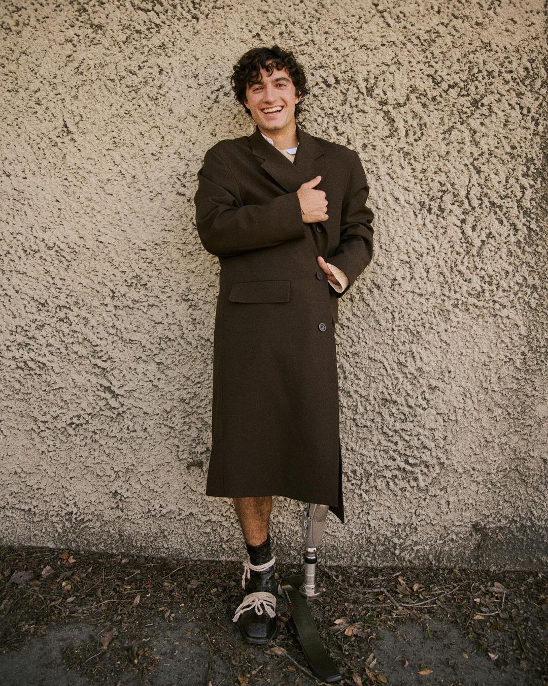A man with a prosthetic leg poses outdoors in a dark brown overcoat and black shoes with white laces, smiling against a textured wall.