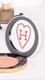 Hermes cosmetic compact with a hand-drawn red heart and letter H on the mirror, with a bottle of perfume in the background.