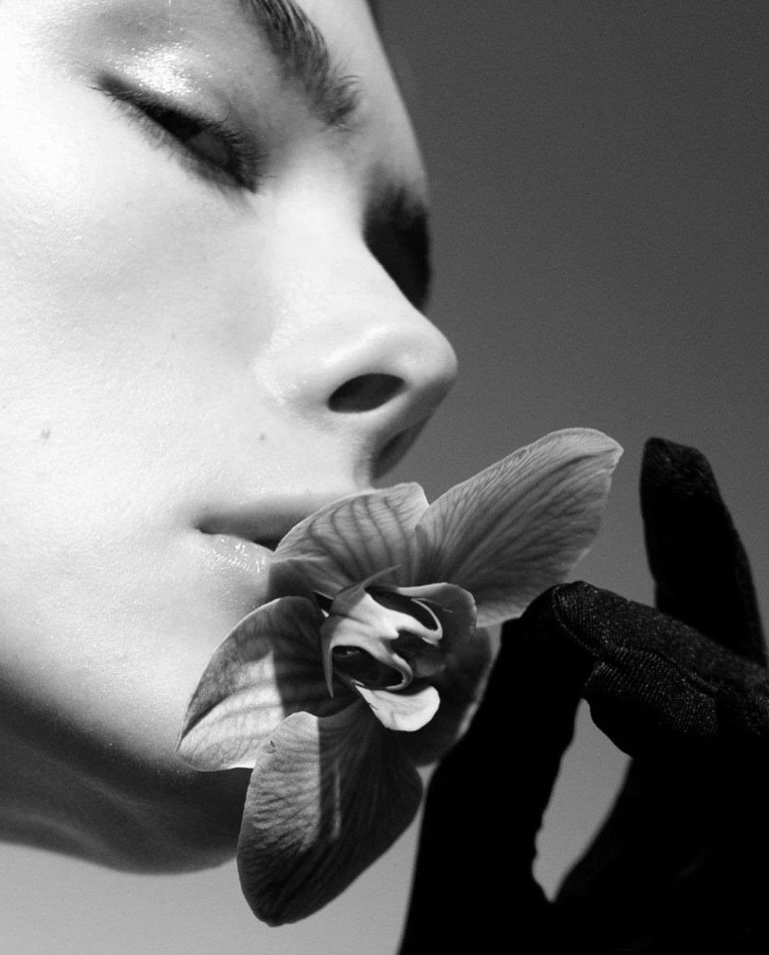 Black and white close-up of a woman's face with closed eyes, holding a delicate orchid flower in a gloved hand, soft lighting.