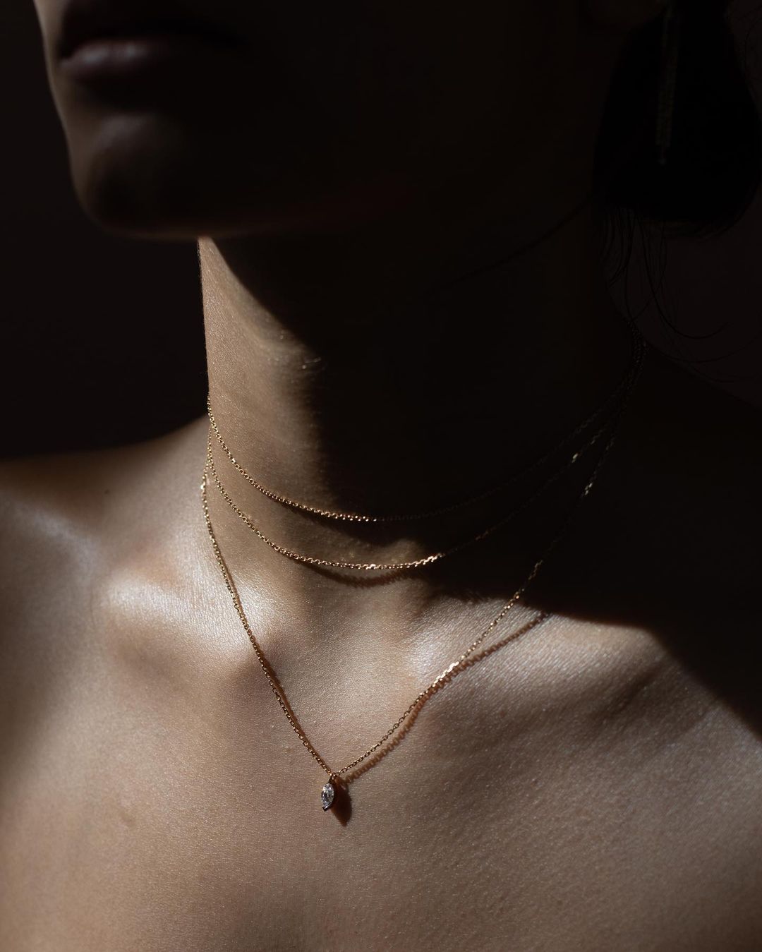 Close-up of a woman's chest and neck with three delicate gold necklaces, softly lit against a dark background, fashion and beauty.