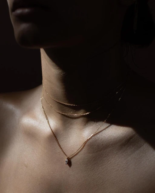 Close-up of a woman's chest and neck with three delicate gold necklaces, softly lit against a dark background, fashion and beauty.
