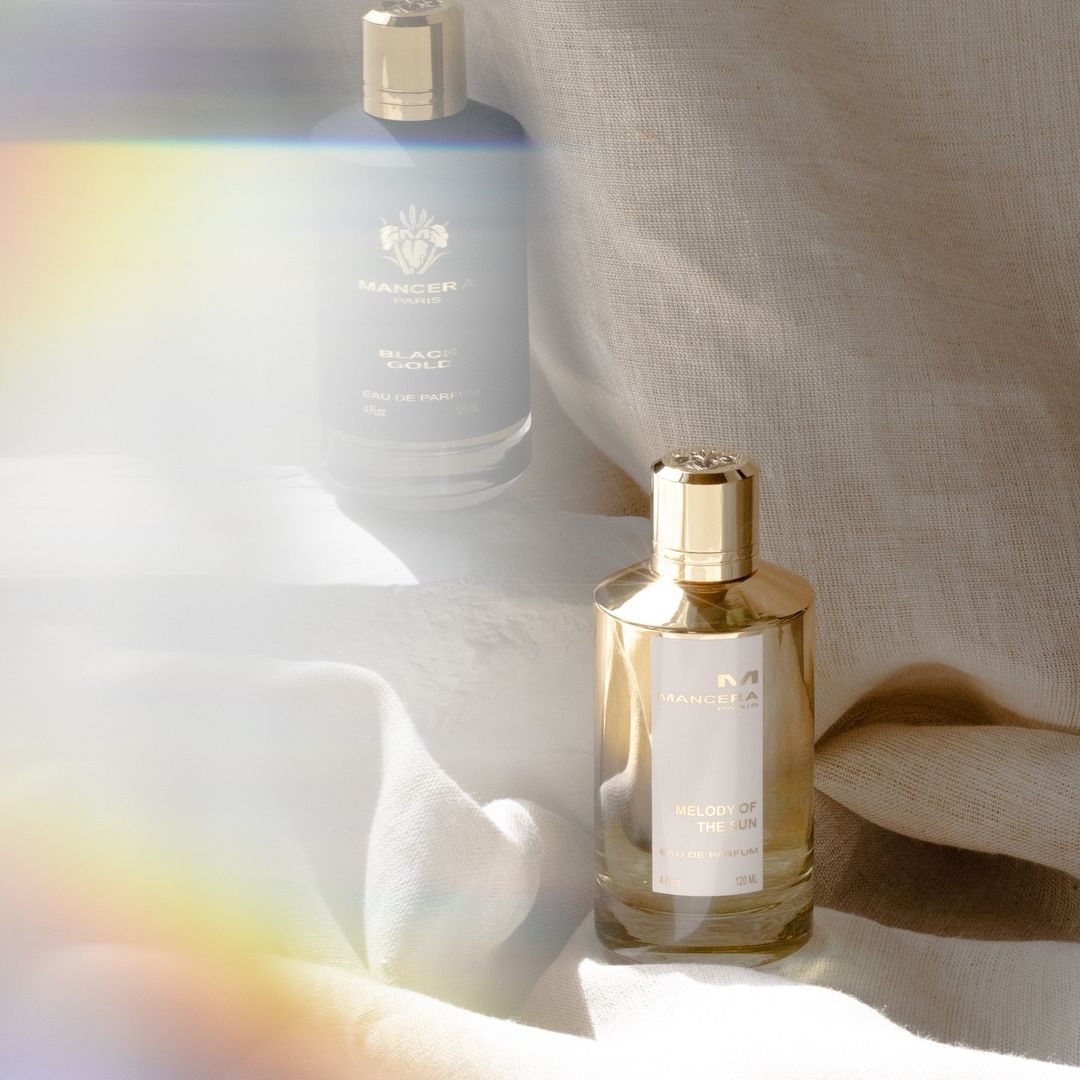 Two Mancera perfume bottles, a gold and black design, are on light fabric against a neutral backdrop with a subtle rainbow lens flare.