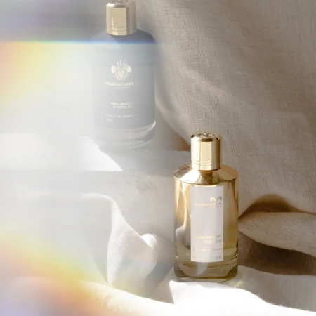 Two Mancera perfume bottles, a gold and black design, are on light fabric against a neutral backdrop with a subtle rainbow lens flare.