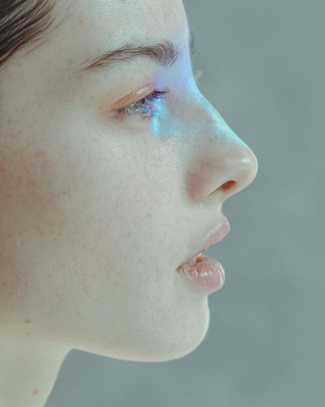 Close-up side view of a fair-skinned adult woman with freckles, blue light on her eye, and neutral expression in a studio setting.