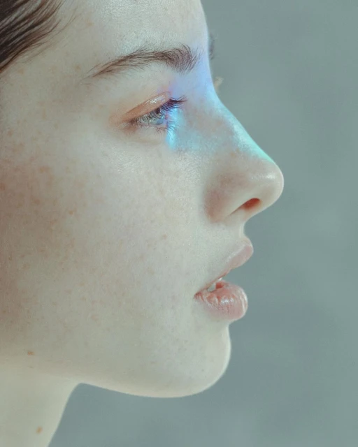 Close-up side view of a fair-skinned adult woman with freckles, blue light on her eye, and neutral expression in a studio setting.