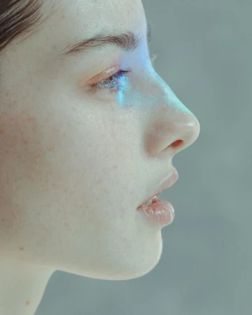 Close-up side view of a fair-skinned adult woman with freckles, blue light on her eye, and neutral expression in a studio setting.
