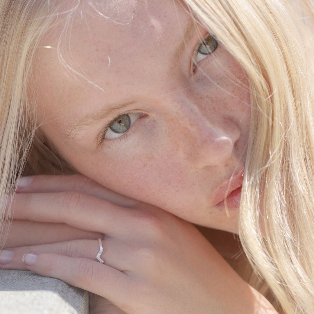 Close-up portrait of a young adult woman with blonde hair and freckled skin, wearing a silver ring, illuminated by soft natural light.