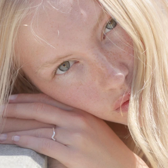 Close-up portrait of a young adult woman with blonde hair and freckled skin, wearing a silver ring, illuminated by soft natural light.