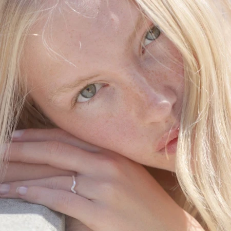 Close-up portrait of a young adult woman with blonde hair and freckled skin, wearing a silver ring, illuminated by soft natural light.
