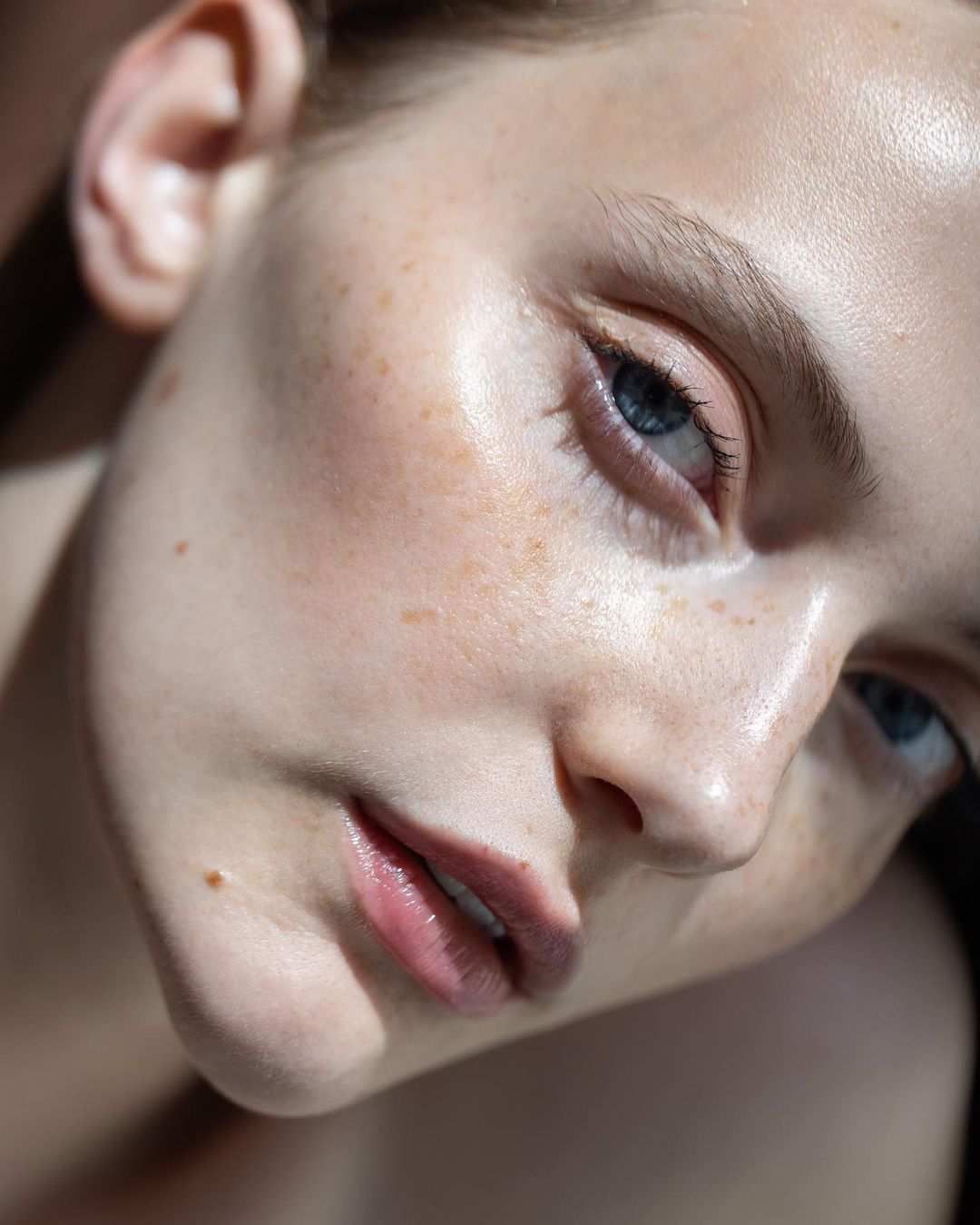 Close-up portrait of a young adult female model with light skin, freckles, and blue eyes, in soft lighting showcasing beauty and skin texture.