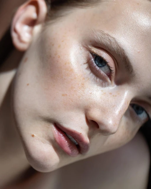 Close-up portrait of a young adult female model with light skin, freckles, and blue eyes, in soft lighting showcasing beauty and skin texture.