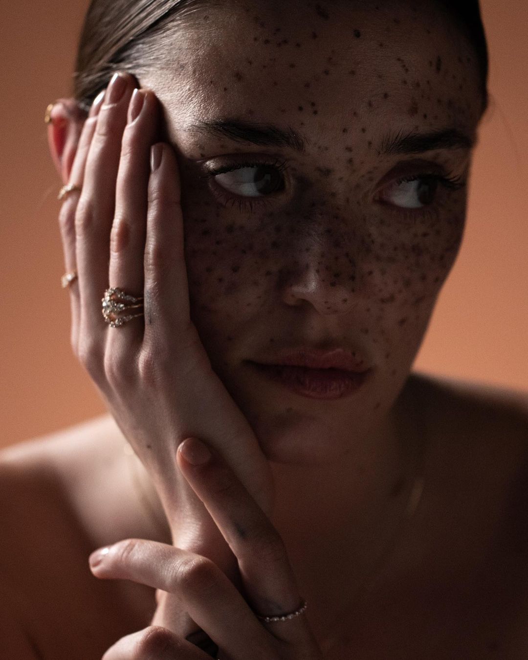 Close-up portrait of a young woman with freckles posing with hand on face, wearing rings, against a neutral orange background.