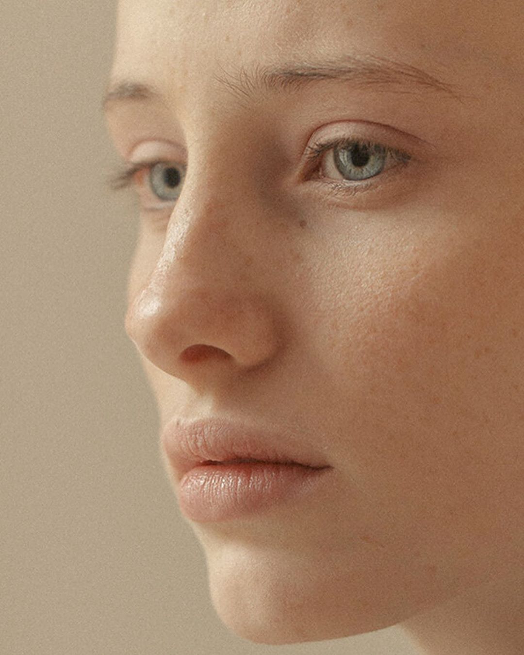 Close-up portrait of a fair-skinned young adult woman with blue eyes and freckles, captured in soft, neutral lighting against a plain background.