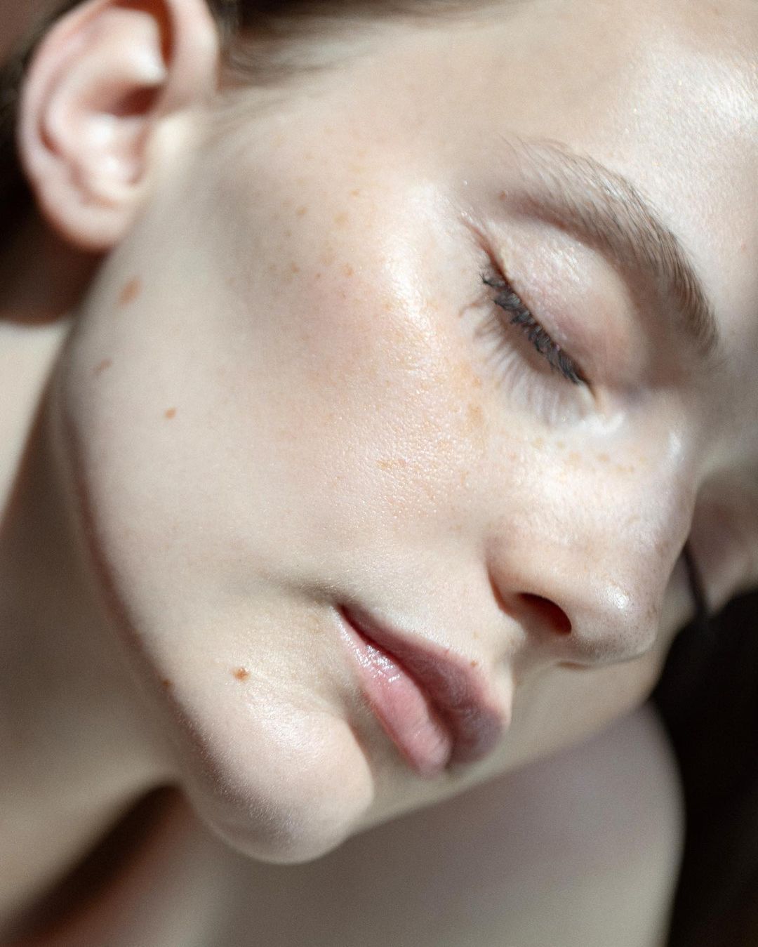 Close-up portrait of a woman's face with freckles, eyes closed, soft lighting, and a dark background focusing on beauty and skincare.