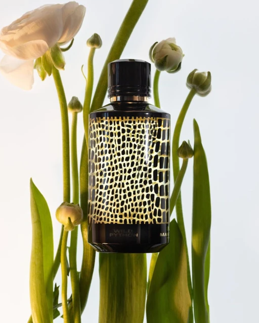 Luxury perfume bottle with a black cap and gold reptile pattern, surrounded by white flowers and green stems against a white backdrop.