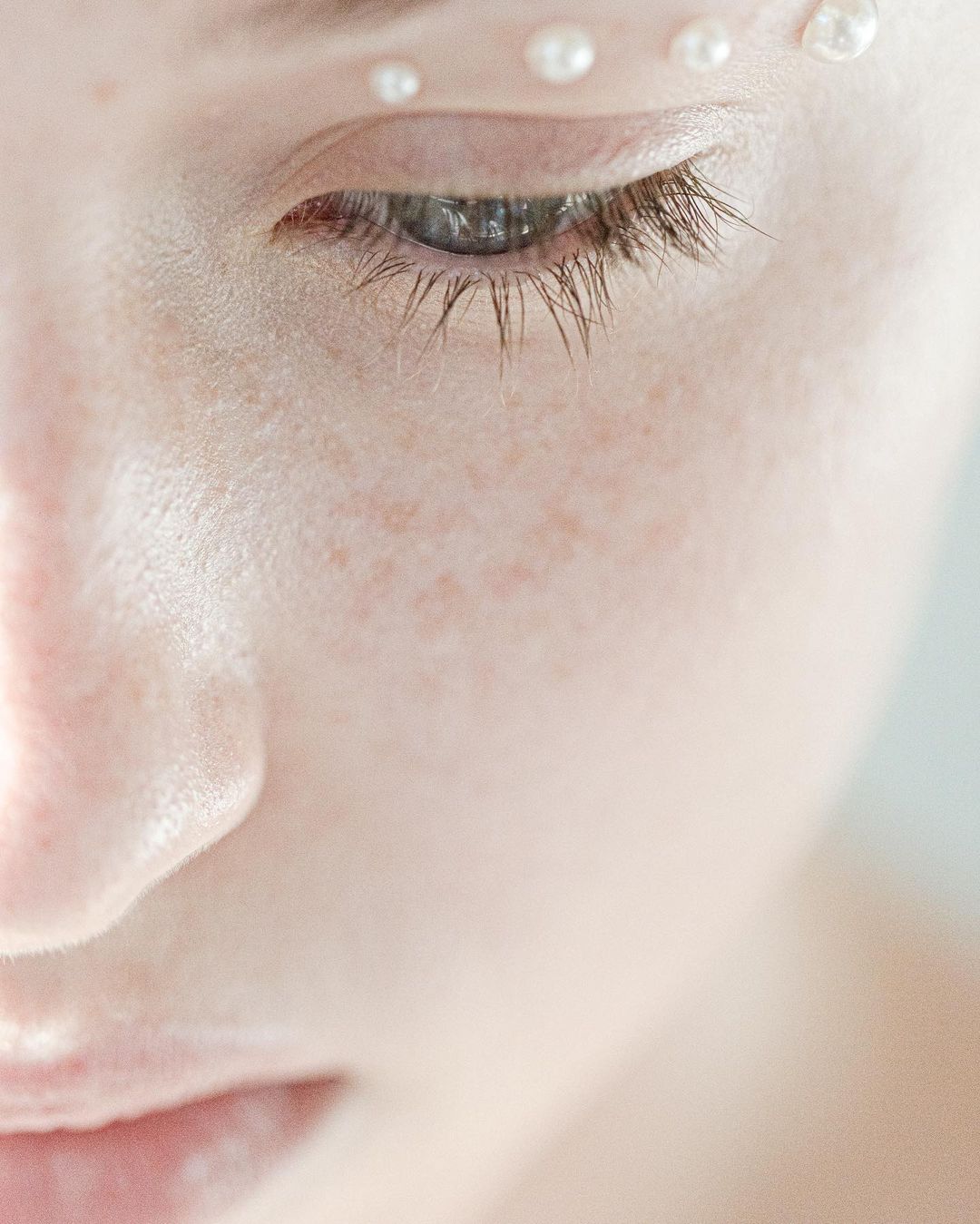 Close-up of a woman's face with fair skin, adorned with small pearls above her eye and on her forehead in a beauty or fashion context.