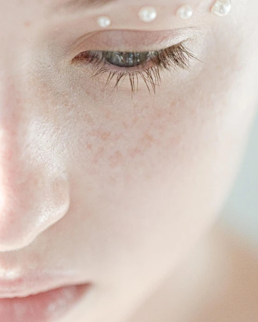 Close-up of a woman's face with fair skin, adorned with small pearls above her eye and on her forehead in a beauty or fashion context.