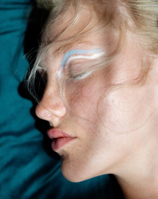 Close-up portrait of a fair-skinned woman with light blonde hair and freckles, lying against a turquoise fabric background. Fashion beauty shot.