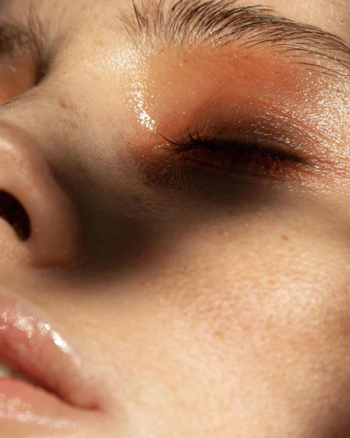 Close-up portrait of a woman's face showing her closed eye, nose, and glossy lips, featuring dewy skin and natural eyebrow hair.