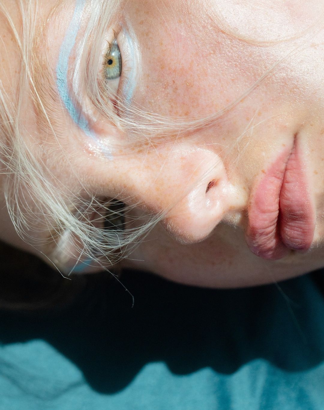 Close-up portrait of a fair-skinned adult female with light blonde hair and blue makeup, lying down on a blue textile surface.