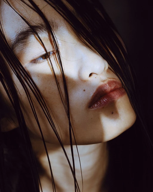Close-up portrait of a person with wet hair partially obscuring the face, highlighting skin texture and dark, glossy lips against a dark backdrop.