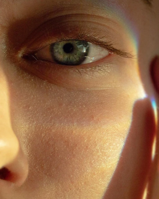 Close-up portrait of a human eye with a light green iris and natural skin texture, with a rainbow prism reflection on the cheek.