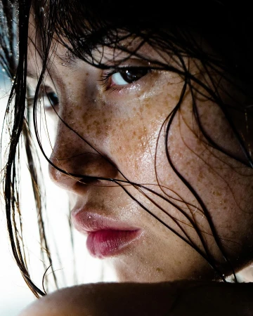 Close-up portrait of a woman with wet skin, dark wet hair strands, and visible freckles shot by Laurent Castellani.