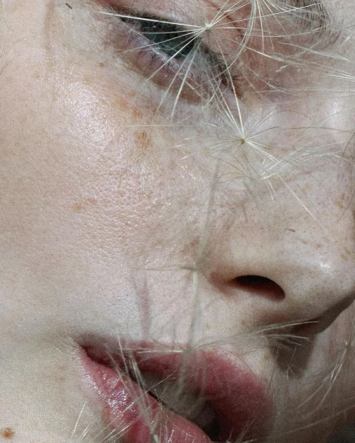 Close-up of an adult woman's face showing her eye, nose, and mouth, with dandelion seed parachutes lightly overlaying her features.
