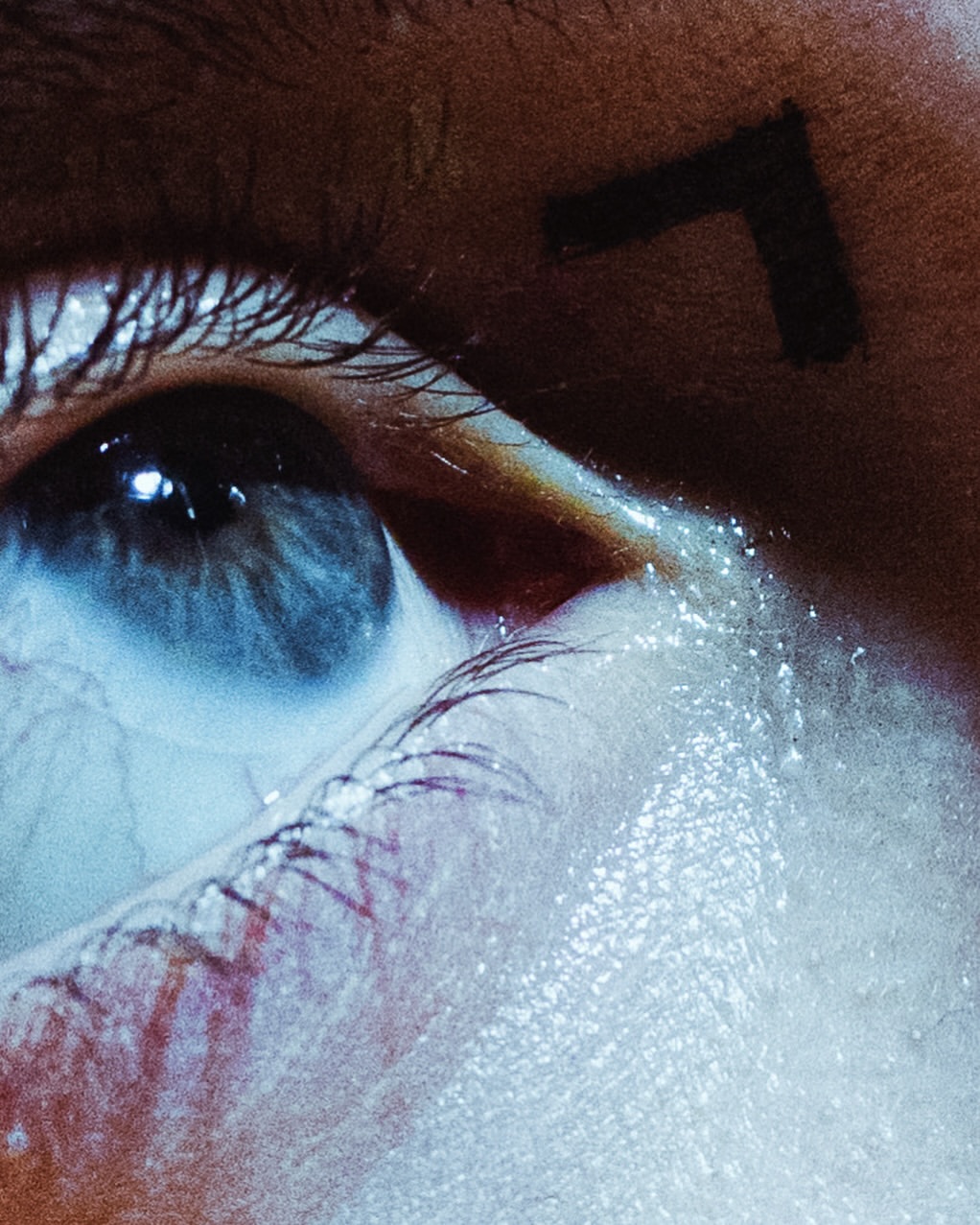 Close-up of a human eye with blue iris, surrounded by eyelashes and skin. There's a dark symbol on the person's forehead.