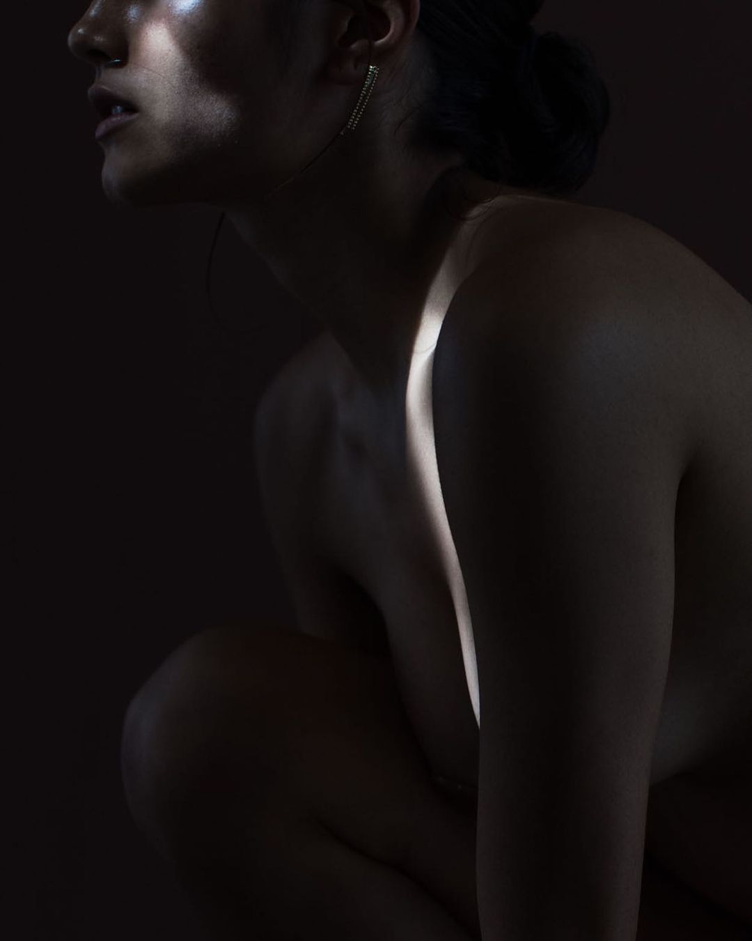Striking portrait of a woman in a crouching pose, dramatically lit to emphasize her form against a dark backdrop, evoking fine art nude.