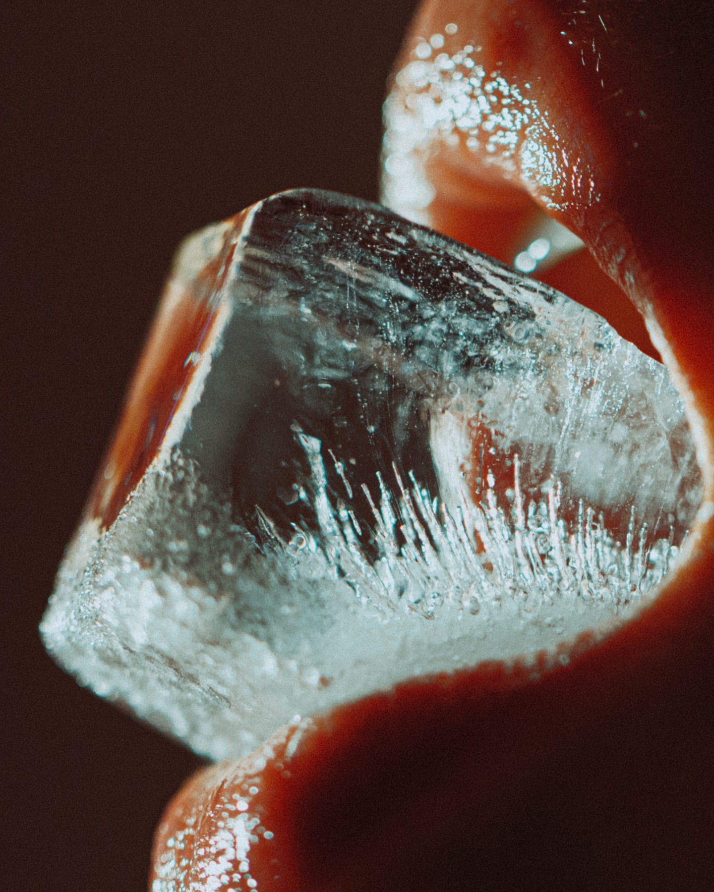 A macro shot highlights a clear ice cube held to glossy lips, emphasizing textures and cool tones against a dark, indistinct background.