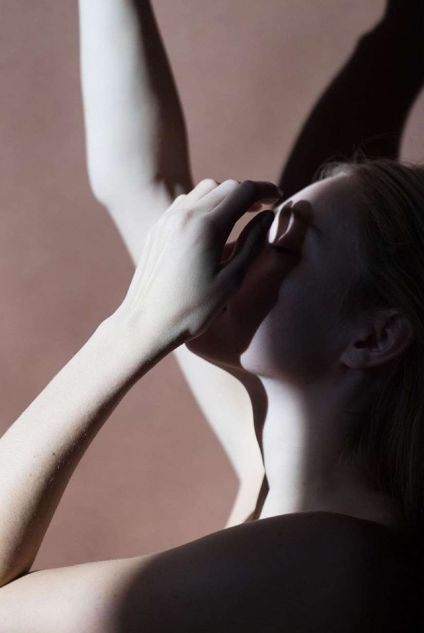 Artistic portrait of fair-skinned woman with hands near her face, partially obscured by shadow. Contrasting light on neutral background.