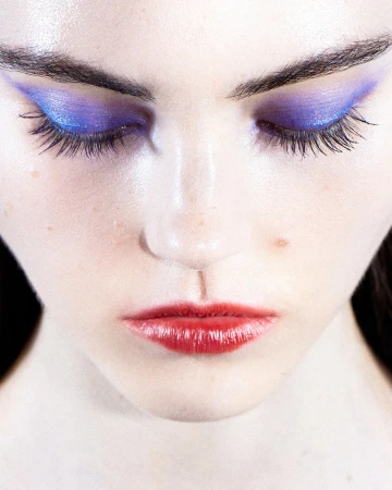 Close-up portrait of a woman with vibrant blue eyeshadow and glossy red lips against a neutral background