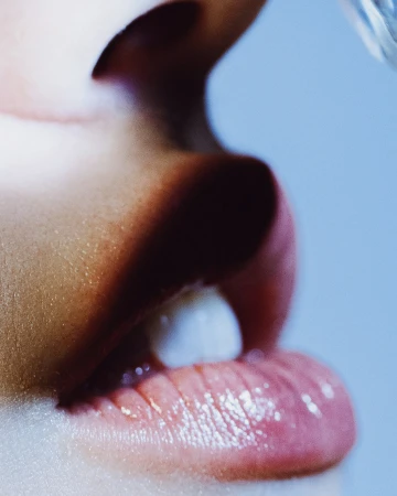 Close-up of a woman's face with lips slightly open, revealing a tooth, with a blurred clear object above, set against a light blue background.