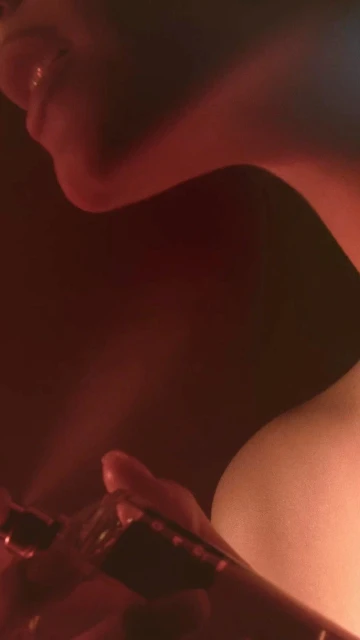 Close-up of a person's neck and shoulder in soft red light with a hand holding a glass bottle, set against a dark background.