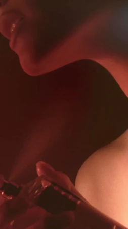 Close-up of a person's neck and shoulder in soft red light with a hand holding a glass bottle, set against a dark background.