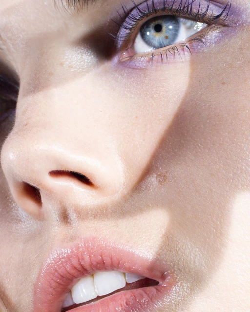 Close-up portrait of a fair-skinned woman with blue eyes and lavender makeup on the upper lids, highlighted by natural, soft lighting.