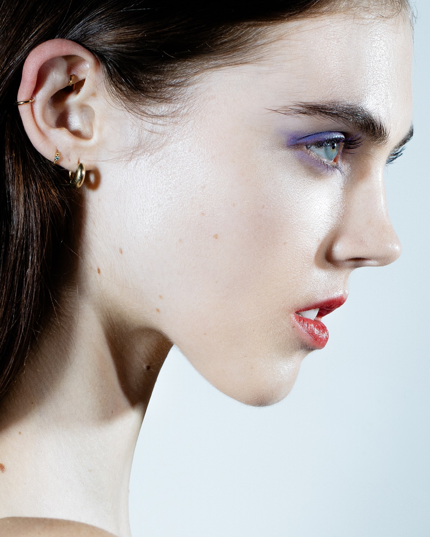 Close-up profile portrait of a young adult female with bright eye makeup and lipstick in a brightly lit studio setting against a light background.