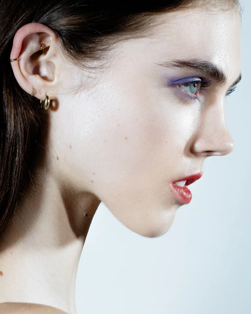 Close-up profile portrait of a young adult female with bright eye makeup and lipstick in a brightly lit studio setting against a light background.