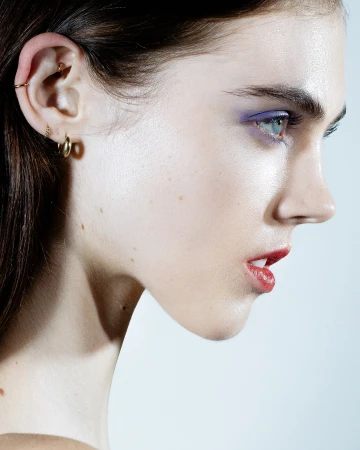Close-up profile portrait of a young adult female with bright eye makeup and lipstick in a brightly lit studio setting against a light background.