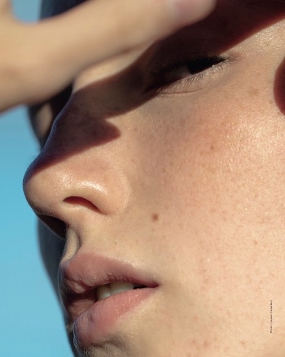 Close-up of a young adult's face against a blue backdrop; hand shields the eyes from sunlight, creating contrasting shadows.